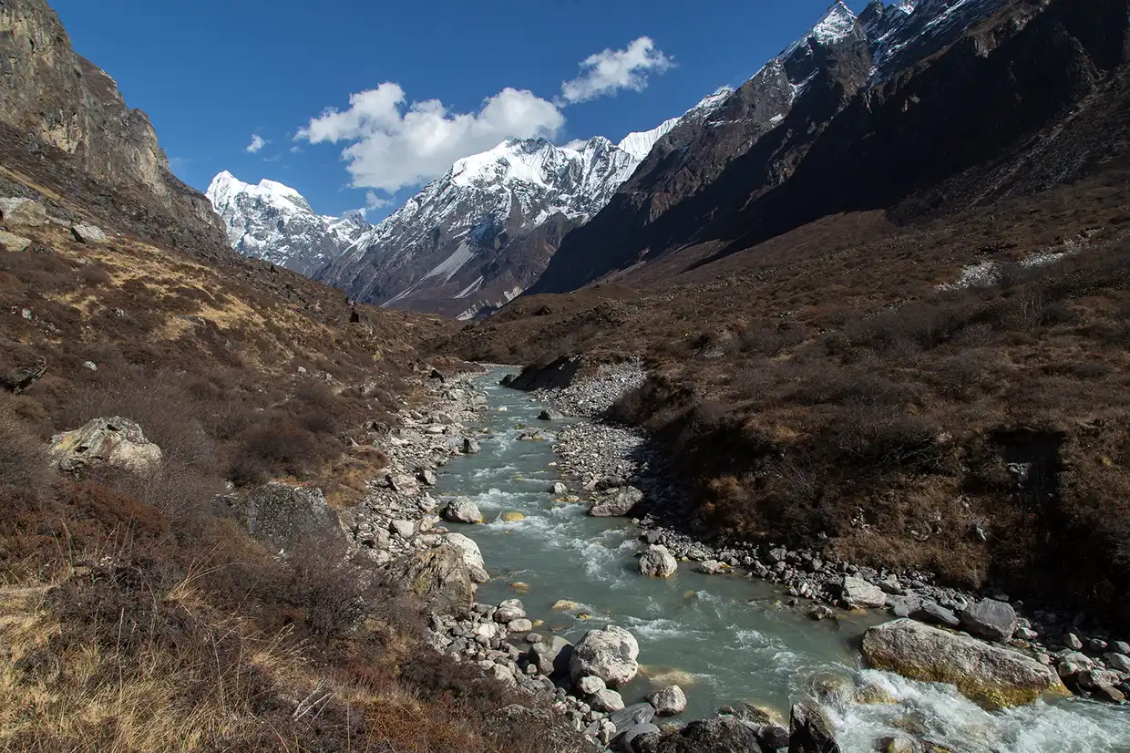 A glacier-fed river flows through a rocky valley beneath towering snow-capped peaks, a scenic landscape highlighted in our Langtang Gosaikunda Trek Guide.