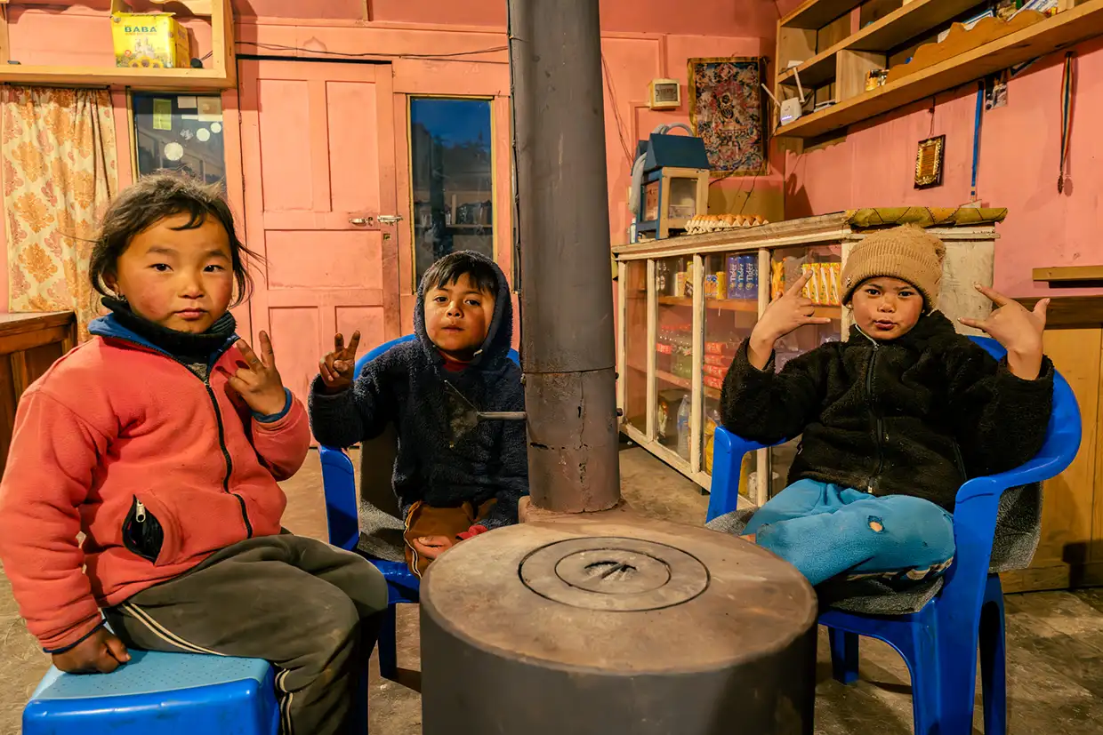 Three young local children wear warm winter clothes and smile while sitting around a central wood stove inside a cozy mountain lodge during a Pikey Peak Trek.