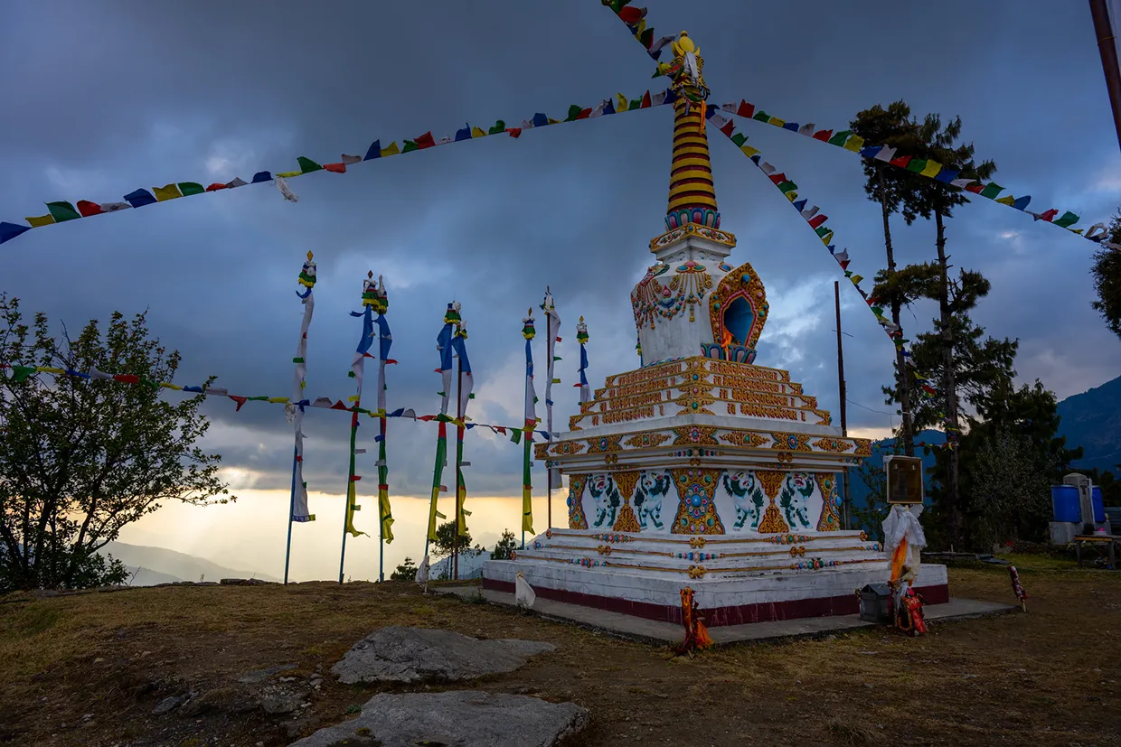 A colorful Buddhist stupa draped in prayer flags stands on a grassy ridge at sunset, a serene cultural landmark along a Langtang Trek Itinerary.