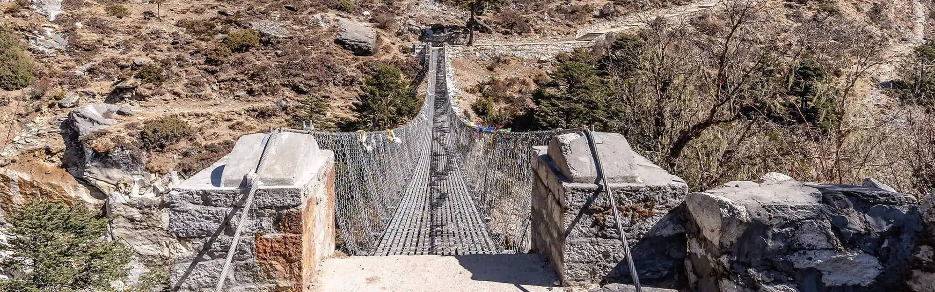 A sturdy metal suspension bridge stretches across a deep, rocky ravine on a sunny day. It is an iconic feature when Hiking in Nepal by Month.