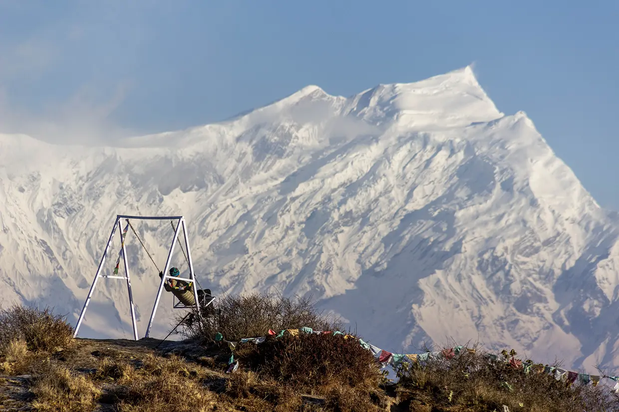 A traveler sits on a scenic overlook swing facing a massive, snow-covered Himalayan mountain peak during the adventurous Khopra Ridge Trek.