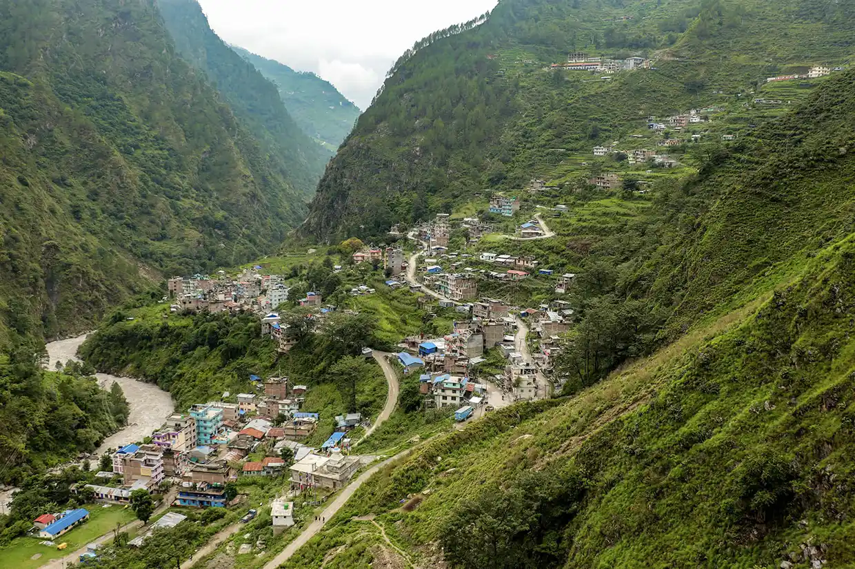 A view looking down onto a vibrant, small village nestled in a lush green mountain valley with a winding dirt road and a river, typical of the stunning landscapes experienced on a Langtang Valley Trek.