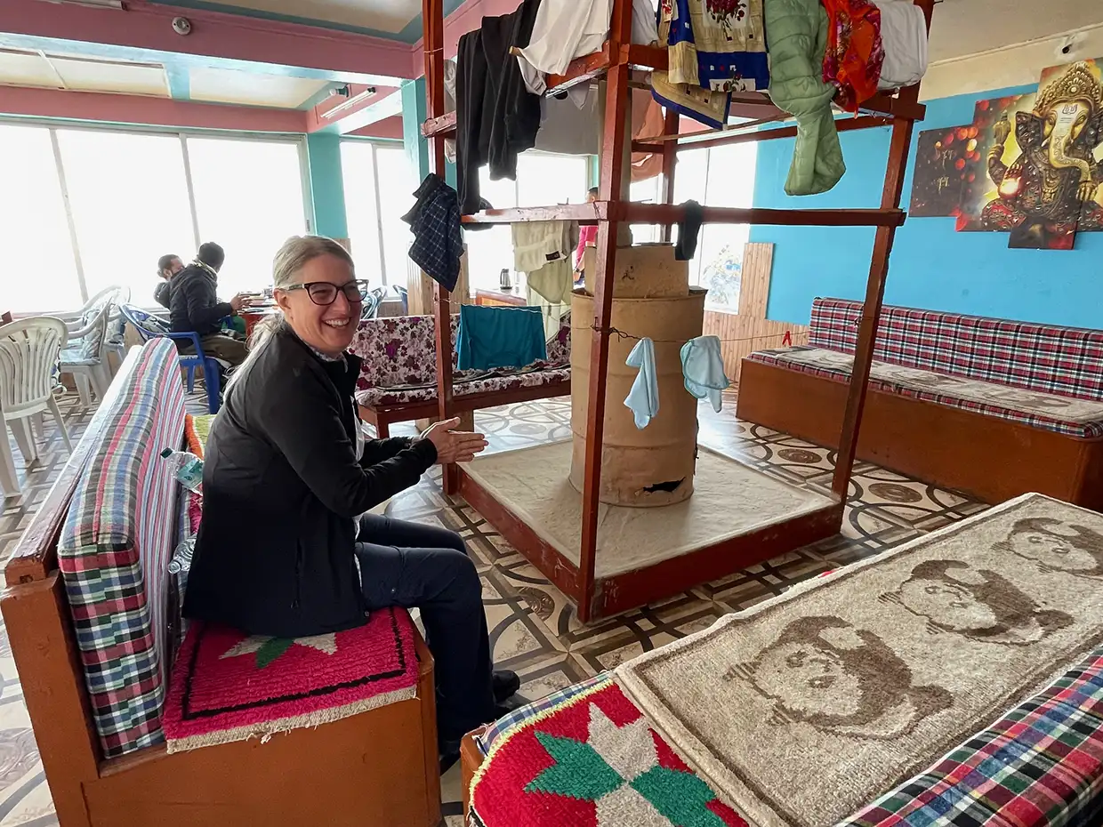 A smiling woman relaxes on a colorful bench near a central warming stove inside a traditional Nepalese tea house during the Khopra Ridge Trek.