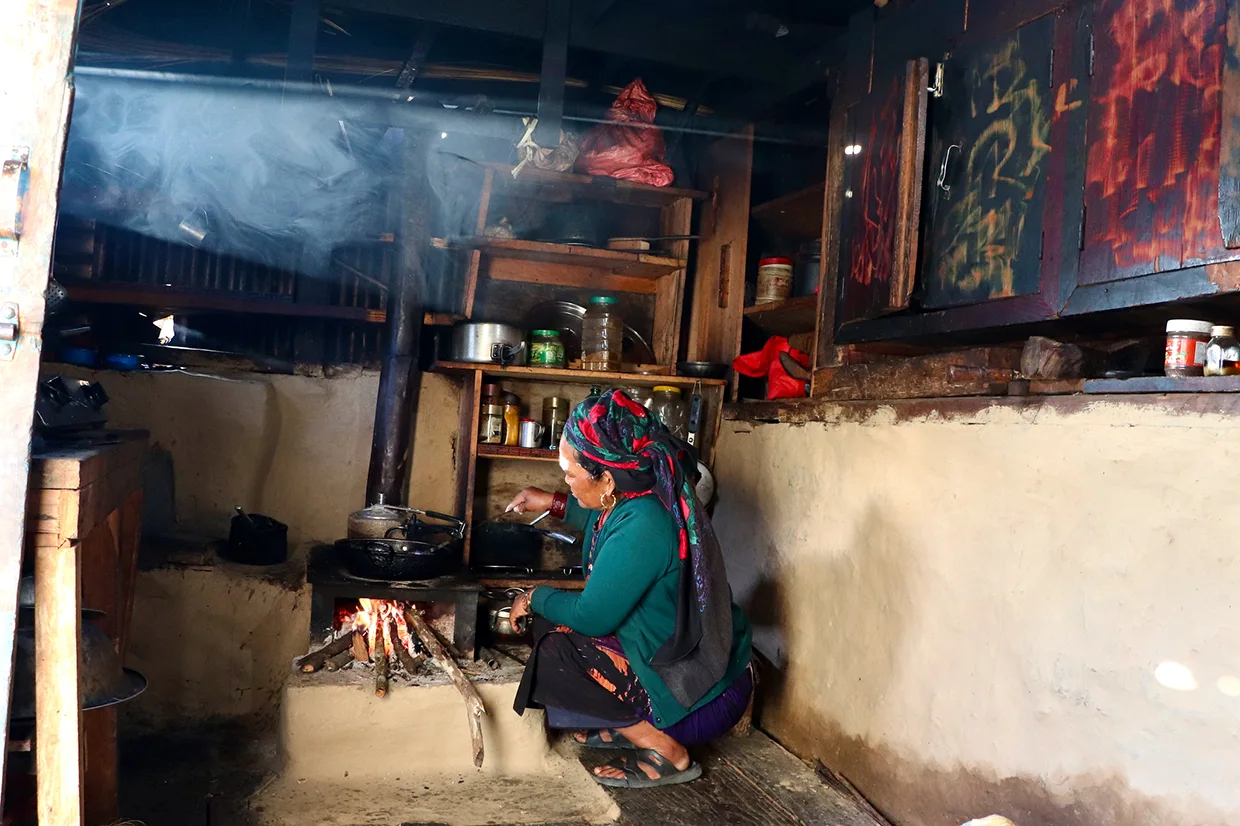 A woman cooks over an open wood fire inside a rustic mud-walled teahouse kitchen. Choosing between the Manaslu vs Annapurna Circuit highlights major differences in lodging comfort.