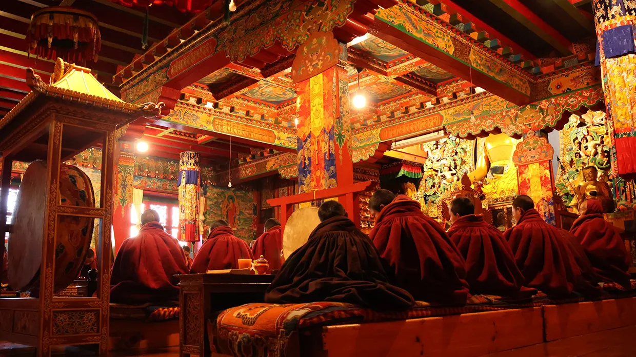 Buddhist monks in red robes sit in prayer inside a colorful, ornate monastery during an Everest Panorama Trek Guide expedition. The room features detailed traditional architecture.