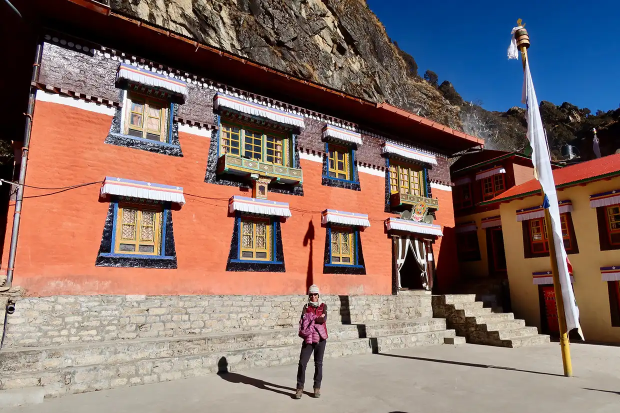 A hiker stands before the vibrant orange walls of Thame Monastery beneath a rocky cliff, a scenic cultural stop on an Everest Cultural Trek.