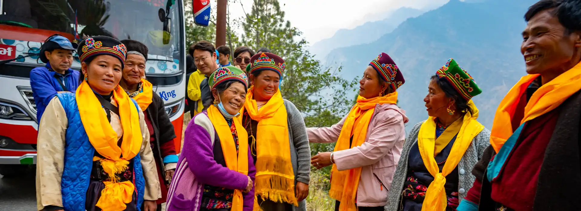 Local villagers dressed in traditional clothing and bright yellow scarves chat near a road on a Tamang Heritage Trek, with a tour bus behind them.