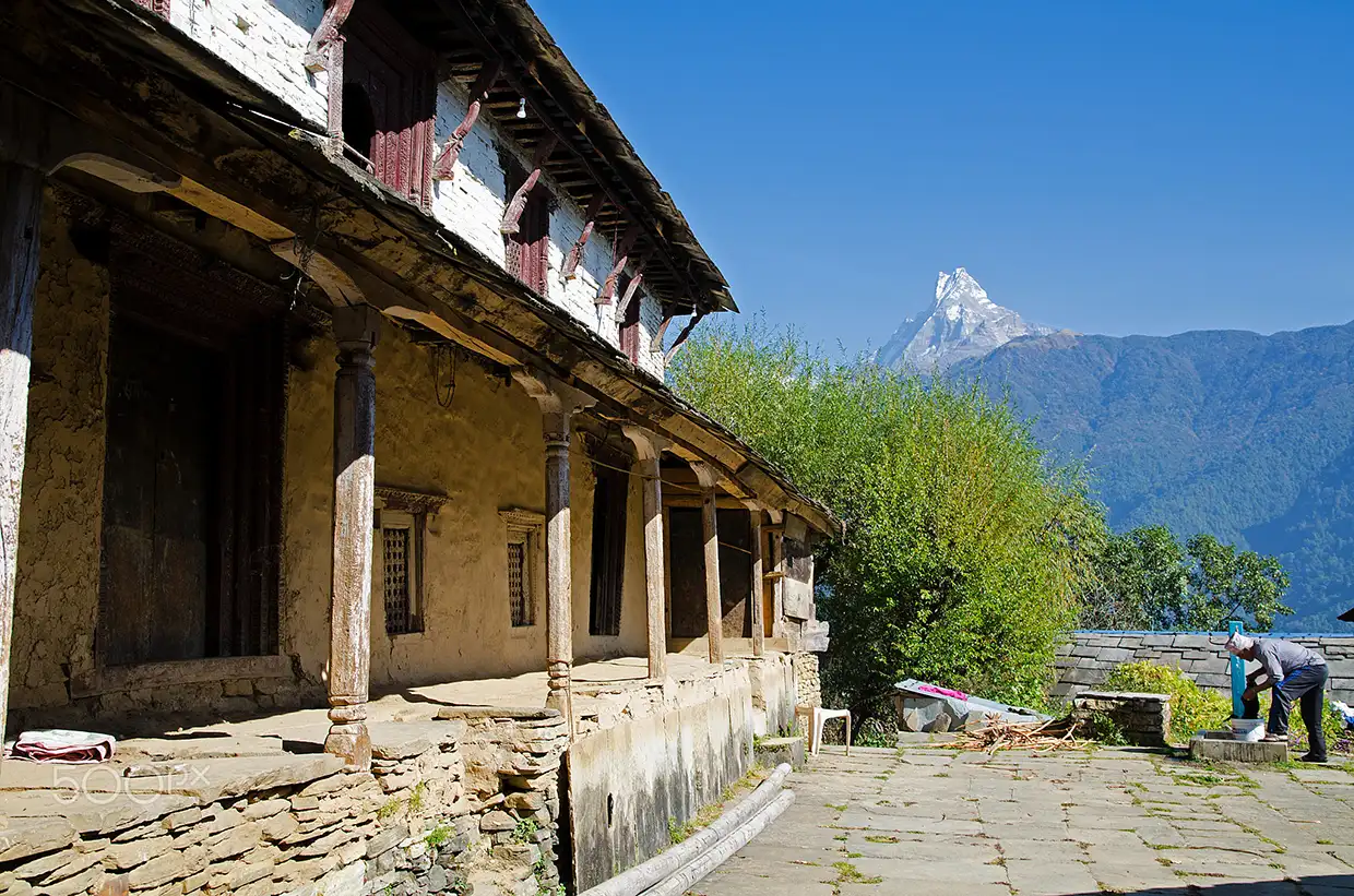 A two-story traditional Gurung house made of stone and clay stands next to a stone path and a small green garden patch during the Annapurna Siklis Trek.