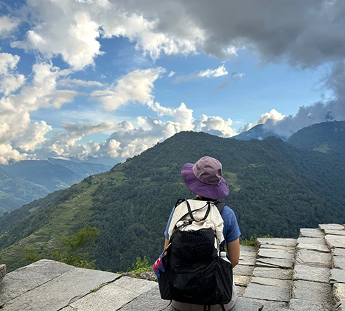 A hiker in a purple sun hat sits on a stone ledge, looking out over lush green mountain valleys and a cloudy blue sky. This Annapurna Circuit Trek Guide helps travelers navigate such scenic trails.