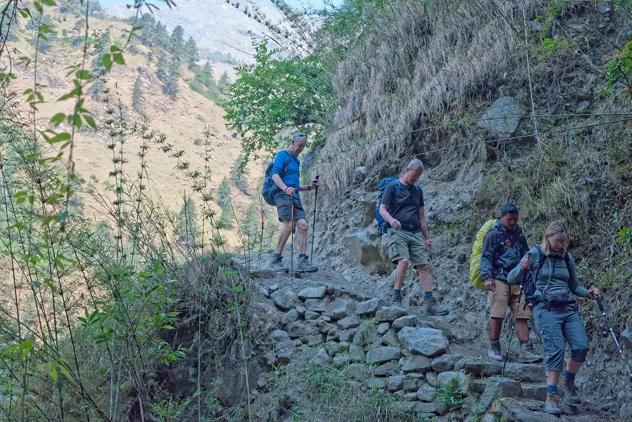 Four hikers with backpacks and trekking poles carefully navigate a narrow, rocky dirt path cut into a steep hillside on a Tamang Heritage Trek.