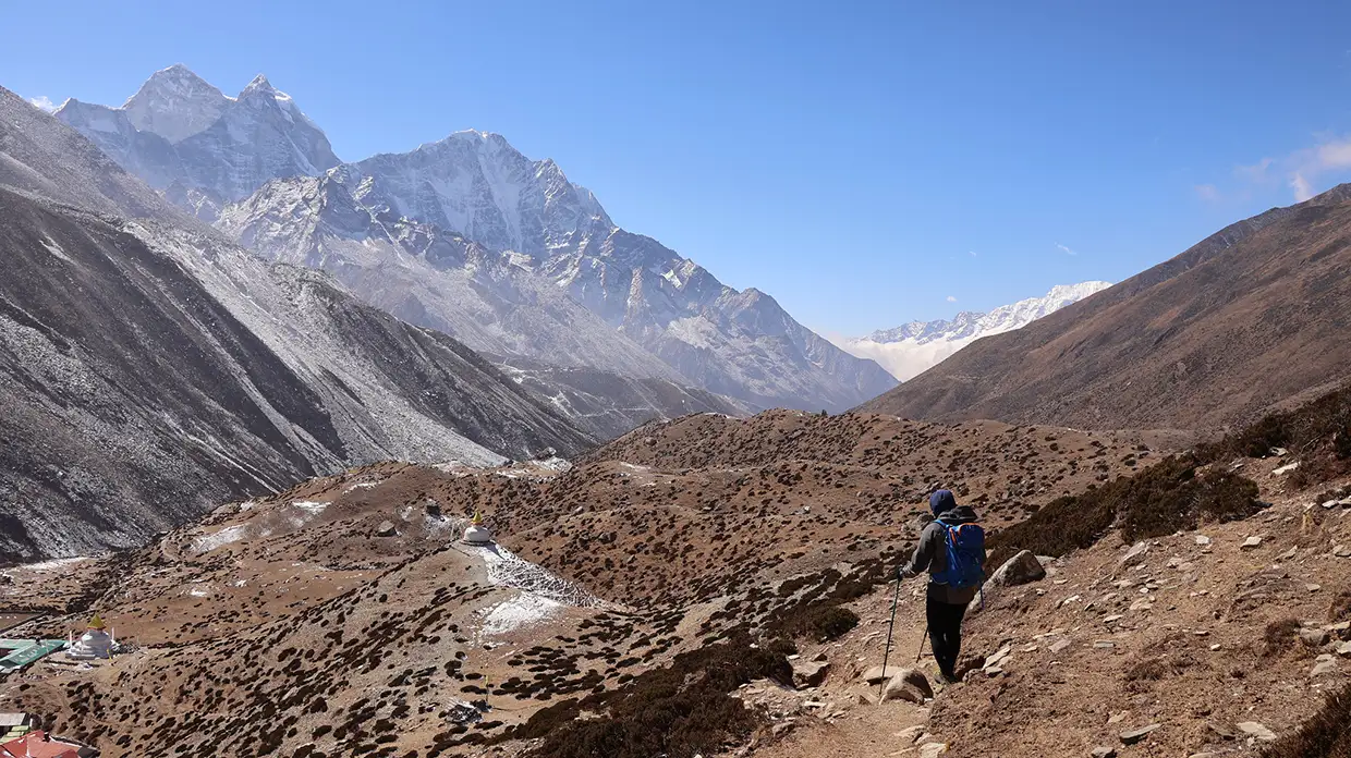 A hiker with a blue backpack walks along a rugged mountain trail toward snow-capped Himalayan peaks. Proper fitness training for Everest Base Camp trek helps trekkers navigate this high-altitude terrain comfortably.