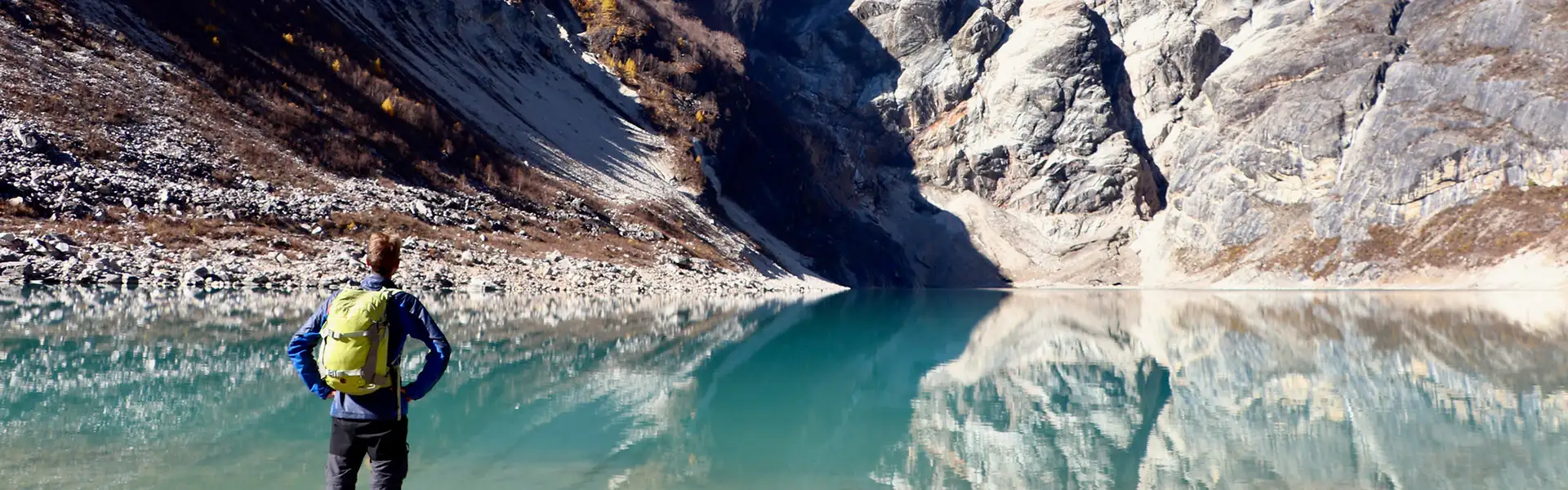 A trekker with a green backpack stands before a calm, turquoise alpine lake. Understanding the Manaslu Circuit Trek Difficulty prepares hikers for these high-altitude rewards.