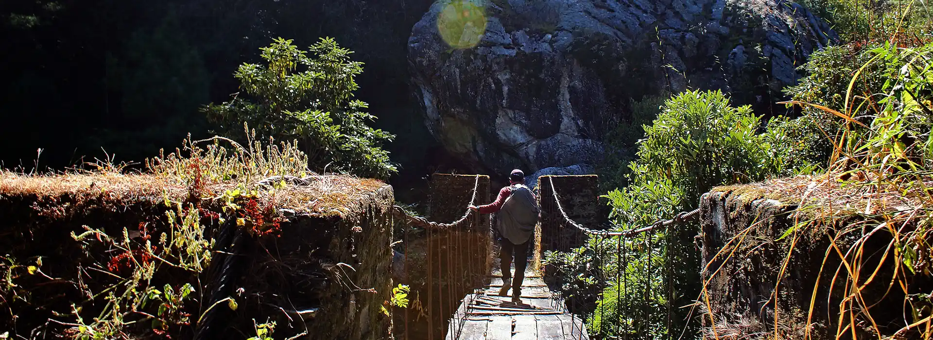 A trekker wearing a large backpack carefully crosses an old, narrow suspension bridge surrounded by lush green foliage and large rocks.