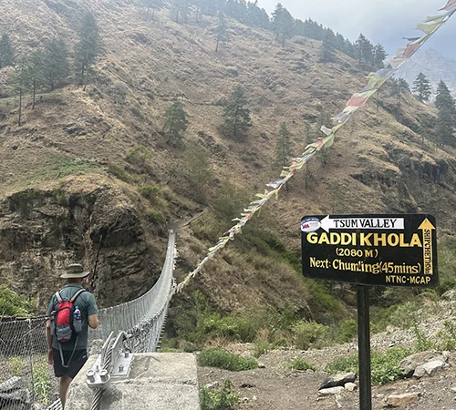 A solo hiker carrying a backpack confidently crosses a suspension bridge over a turquoise river in a mountain gorge, showcasing the best time for the Manaslu Circuit Trek.
