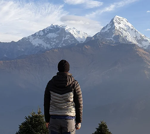 A hiker in a black and grey puffer jacket and beanie, viewed from behind, stands gazing at snow-capped Himalayan mountains. Their warm attire matches a well-planned Annapurna Base Camp Trek Packing List.