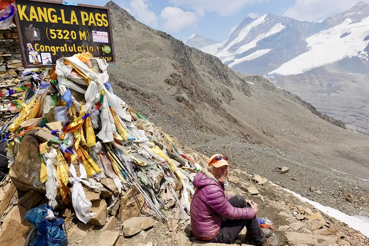 A smiling trekker sits on a rocky slope next to the Kang-La Pass sign and colorful prayer flags during the Nar Phu Valley with Annapurna Circuit Trek.