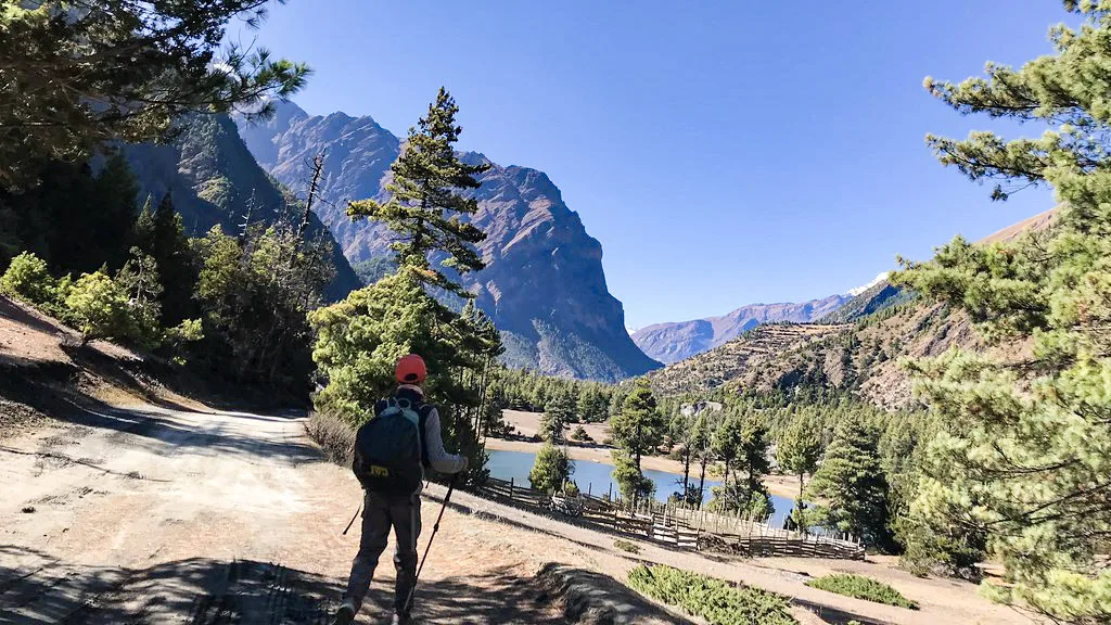 A solo hiker with an orange cap and backpack walks with trekking poles along a dusty mountain trail during the Annapurna Circuit with Tilicho Lake Trek, overlooking a valley.