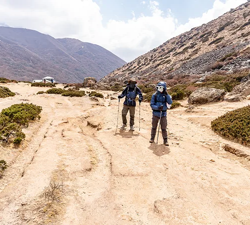 Two trekkers use poles on a dusty mountain path under a bright sky. This describes the varied trail conditions found when Hiking in Nepal by Month.