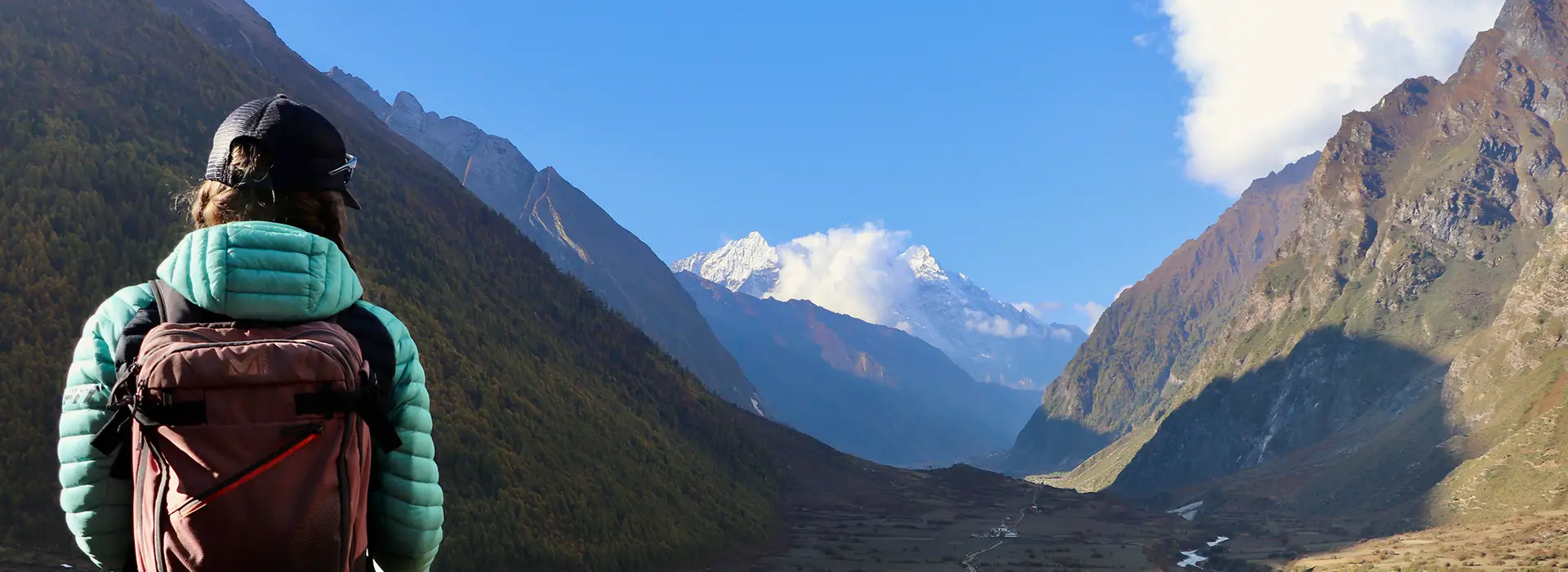 A hiker with a backpack stands on a mountain trail, looking down a vast green valley toward snow-capped peaks, highlighted in our Manaslu Circuit Trek Guide.