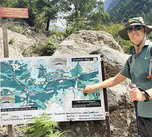 A hiker in a sun hat points to his current position on a trail map board. Gauging the Manaslu Circuit Trek Difficulty helps with route planning.
