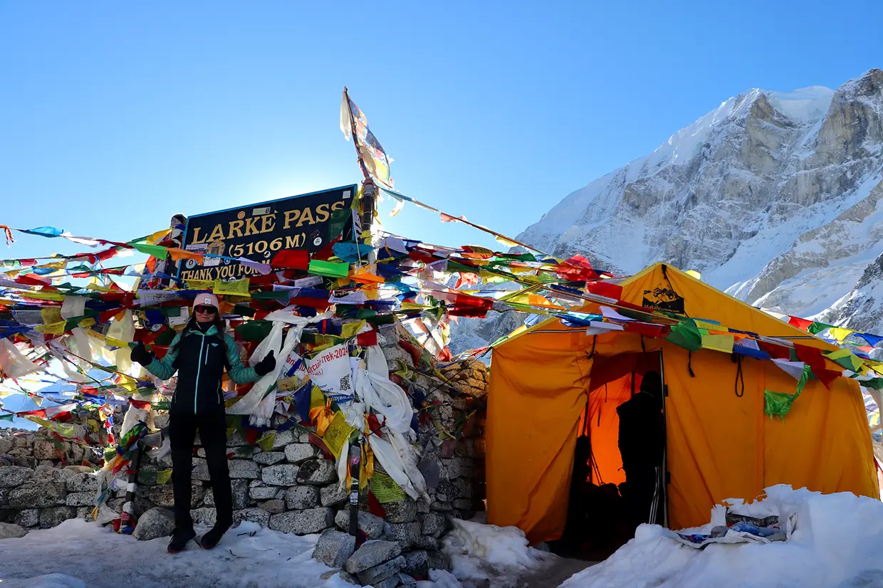 A hiker stands next to the Larke Pass sign at 5106m covered in prayer flags and a yellow tent, ideal for a Manaslu Circuit Trek Guide.