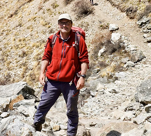 A male trekker in a red fleece jacket and cap stands on a rocky mountain path during the Nar Phu Valley with Annapurna Circuit Trek.