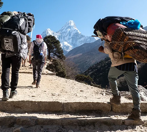 Two trekkers and a local porter carry heavy backpacks up a dirt path toward Ama Dablam mountain on an Everest View Trek.