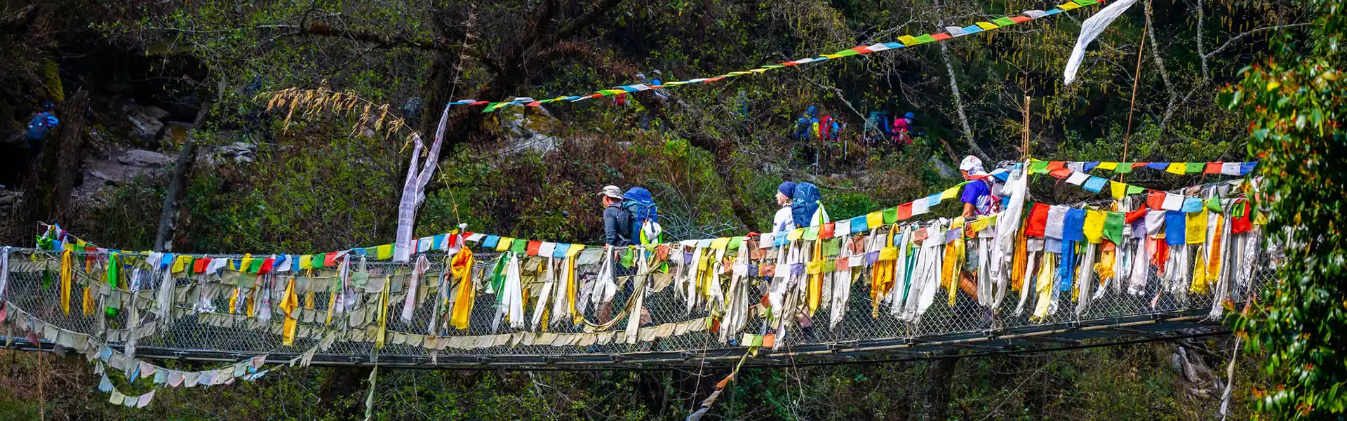 Several trekkers with large backpacks cross a long metal suspension bridge decorated with colorful prayer flags, an iconic experience on a Langtang Trek Itinerary.