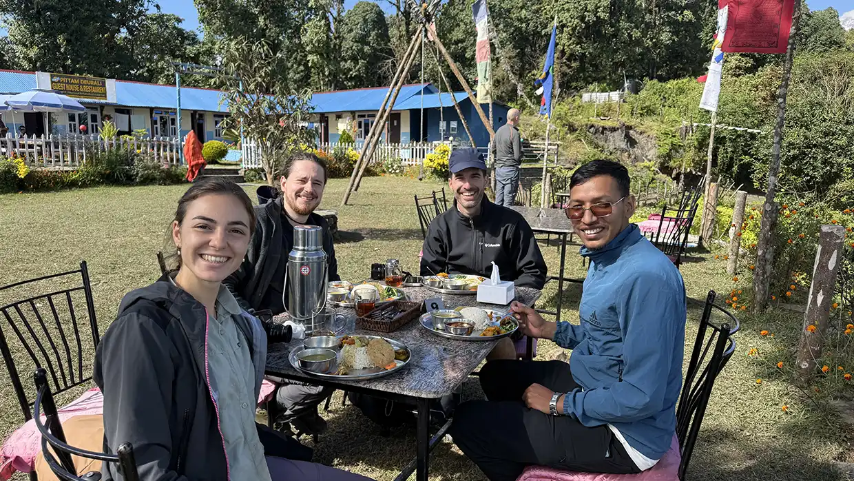 Four smiling travelers share a traditional meal at an outdoor teahouse table under prayer flags. This Mardi Himal Trek Guide highlights the social and cultural aspects of mountain hiking.