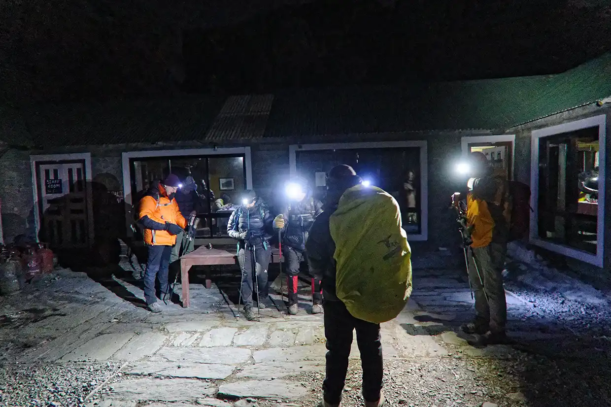 A group of hikers wearing winter gear and bright headlamps gathers outside a stone lodge at night during the Nar Phu Valley with Annapurna Circuit Trek.