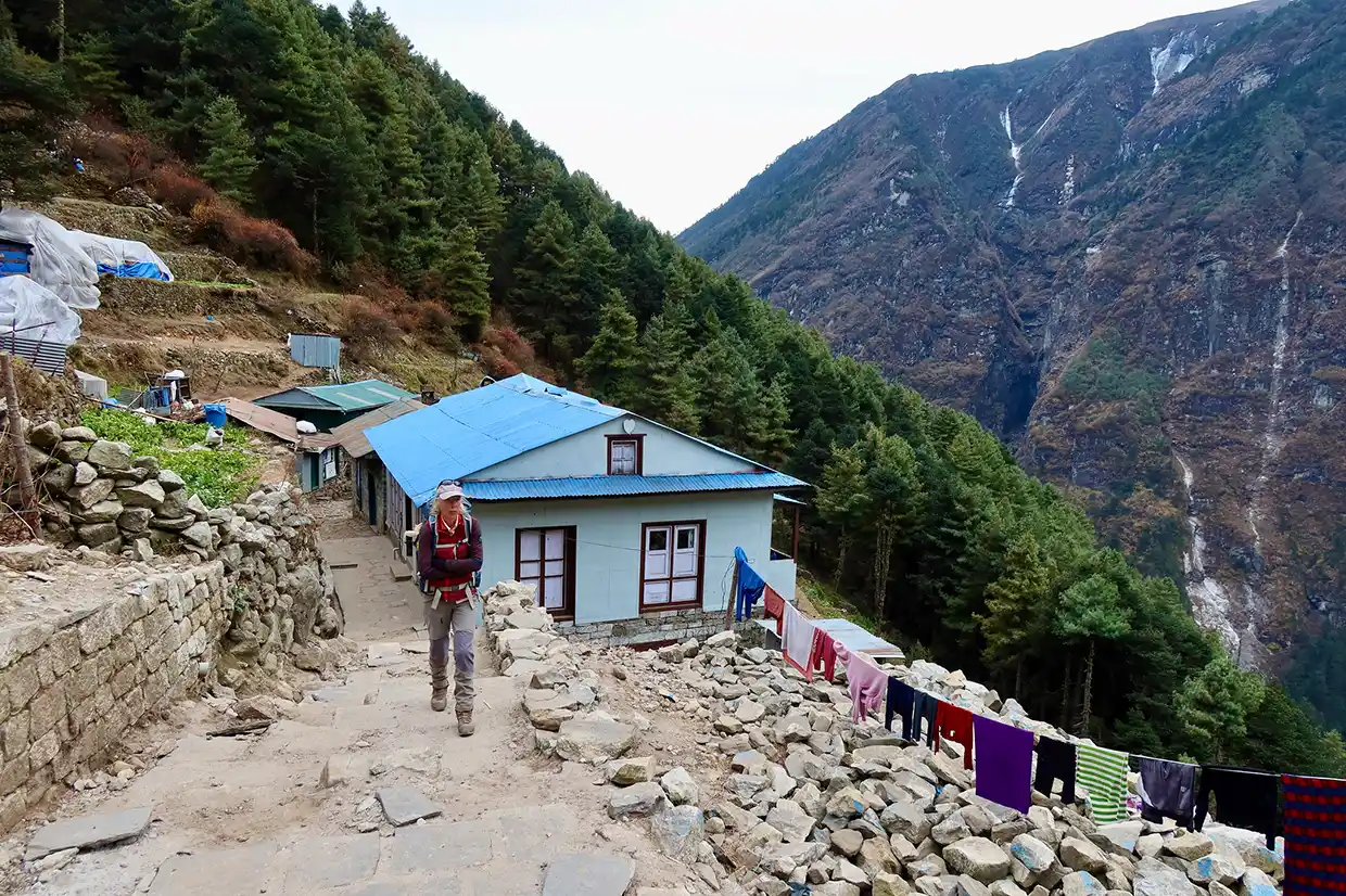 A hiker walks up a stone mountain path past a blue-roofed building, overlooking a steep forested valley during an Everest Cultural Trek.