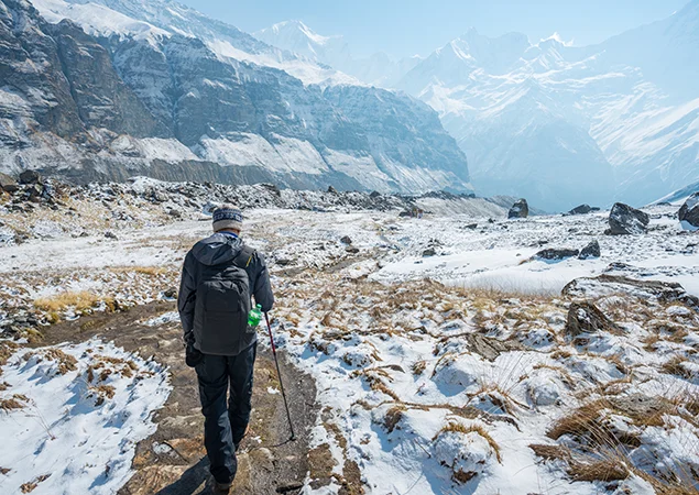 A hiker in winter gear uses a trekking pole to navigate a snowy, rocky trail toward massive white mountain walls during a Thorong La Pass Crossing.