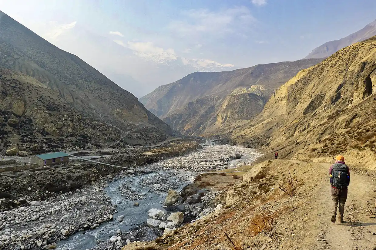 A trekker with a large backpack walks along a wide dirt trail tracking a rocky river through a mountain valley during the Nar Phu Valley with Annapurna Circuit Trek.