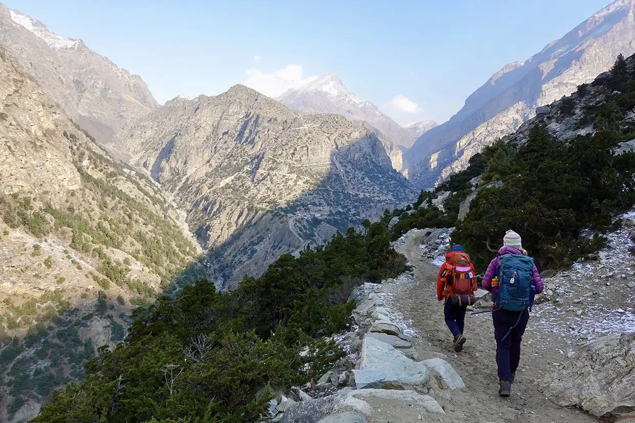 Two hikers with large backpacks walk along a rugged cliffside trail surrounded by massive, rocky canyon walls during the Nar Phu Valley with Annapurna Circuit Trek.