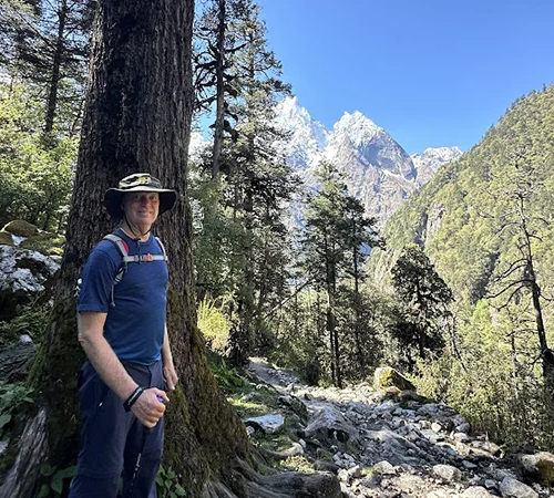 A male trekker stands beside a large tree trunk on a forested mountain path. Our Manaslu Trek Permit Guide assists hikers exploring these remote Nepal valleys.