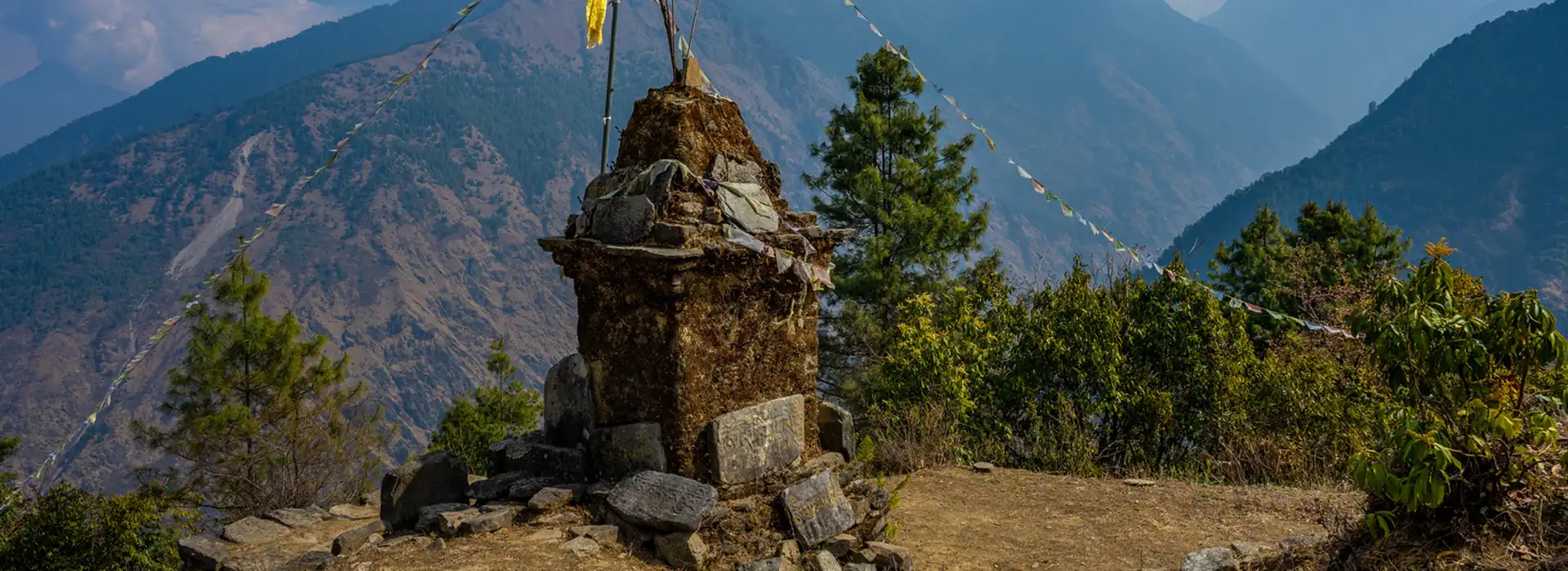 A mossy stone stupa with colorful prayer flags stands on a ridge overlooking a deep valley, a perfect vista for Spring Hiking in Nepal.