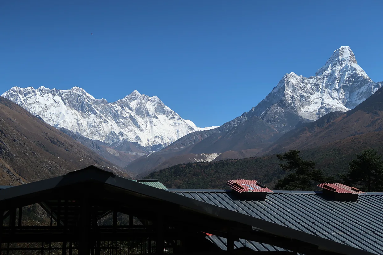 Snowy mountain ridges rise behind dark rooftops, showcasing the towering peak of Ama Dablam under a bright blue sky on an Everest View Trek.