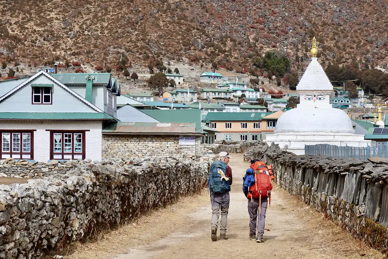 Two hikers with large backpacks walk down a stone path in Khumjung village past a white Buddhist stupa during an Everest Cultural Trek.