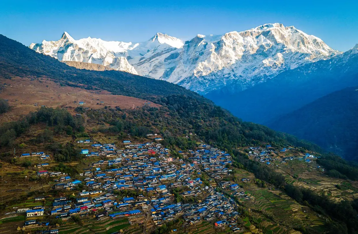 An aerial wide view shows the blue-roofed houses of Sikles village spread across a green hillside under massive snow-covered peaks during the Annapurna Siklis Trek.