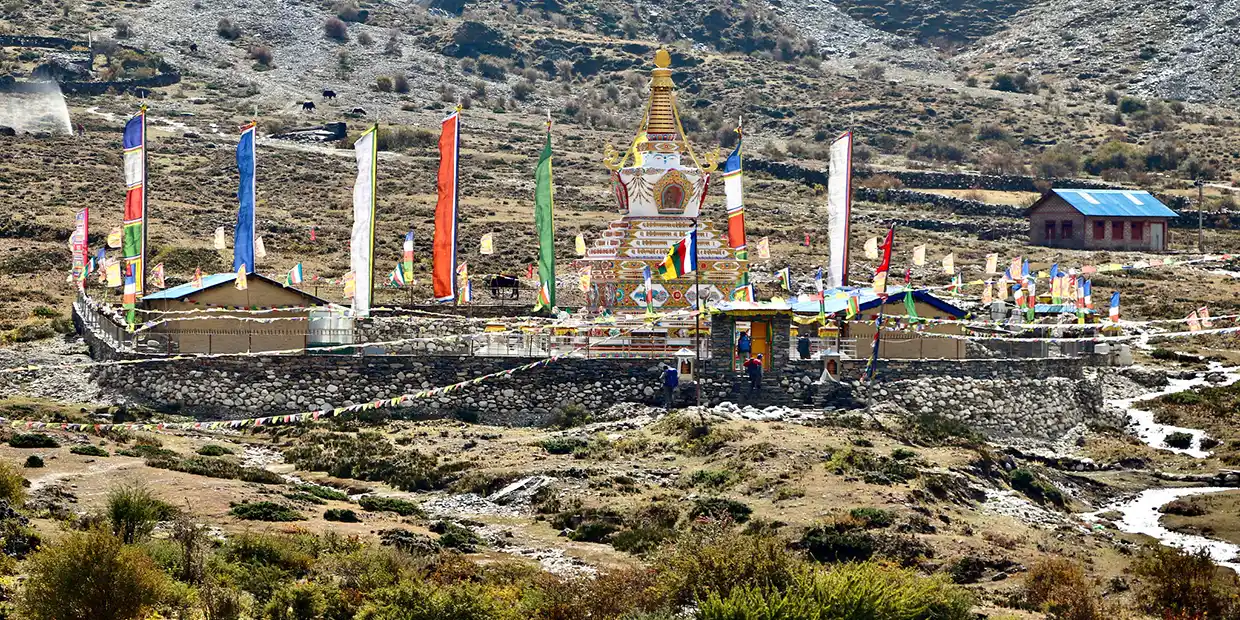 A colorful Buddhist stupa surrounded by prayer flags and a stone wall stands in a high mountain valley during the Manaslu Tsum Valley Trek.