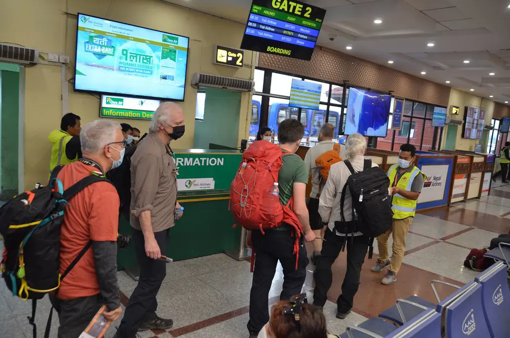 Travelers carrying hiking backpacks stand in line near a boarding gate and flight information screen inside a domestic airport terminal before starting their Upper Dolpo Trek.