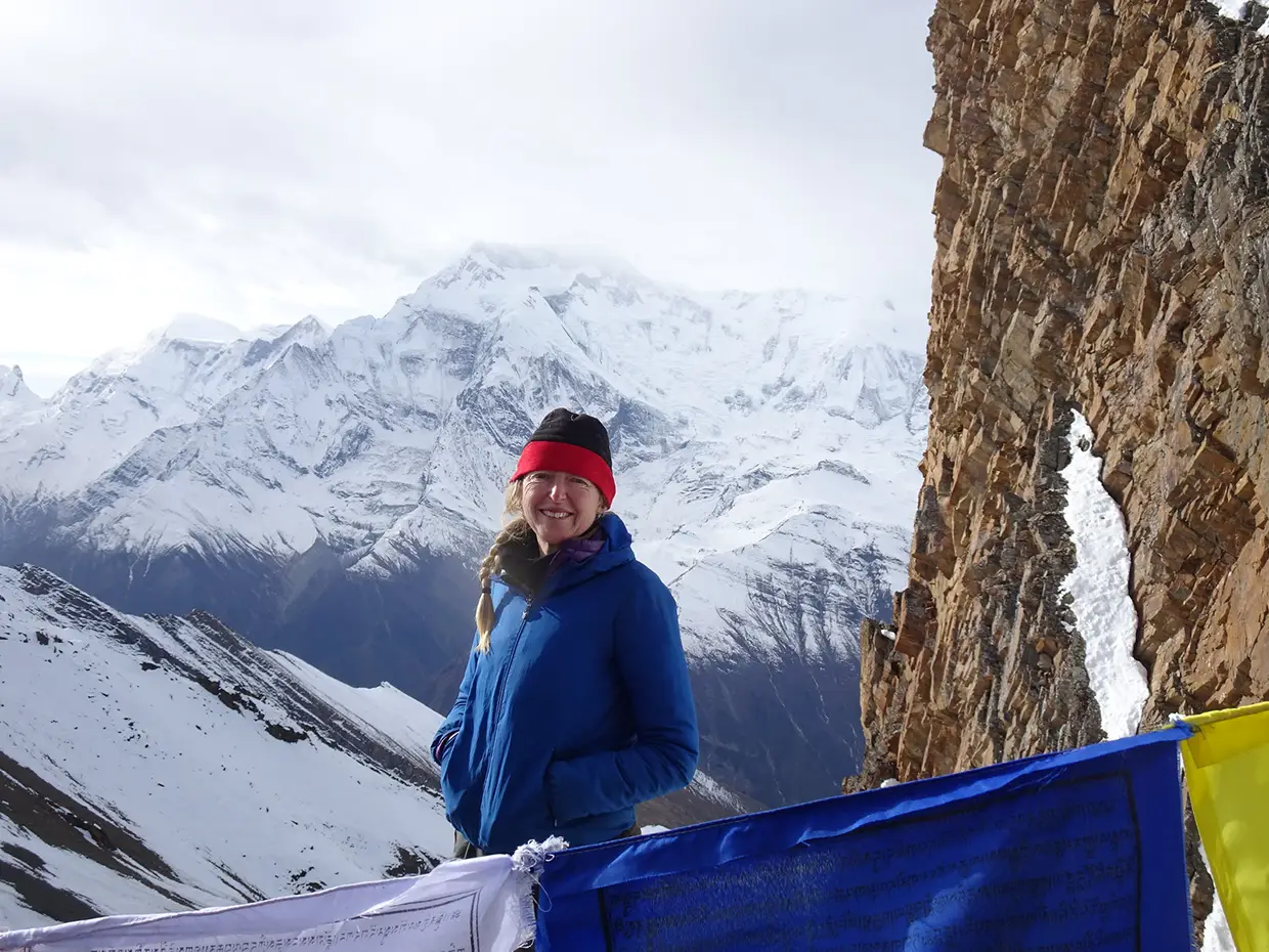 A smiling female hiker in a blue jacket stands near a rocky cliff and colorful prayer flags, with snow-covered mountain peaks behind her during the Upper Dolpo Trek.