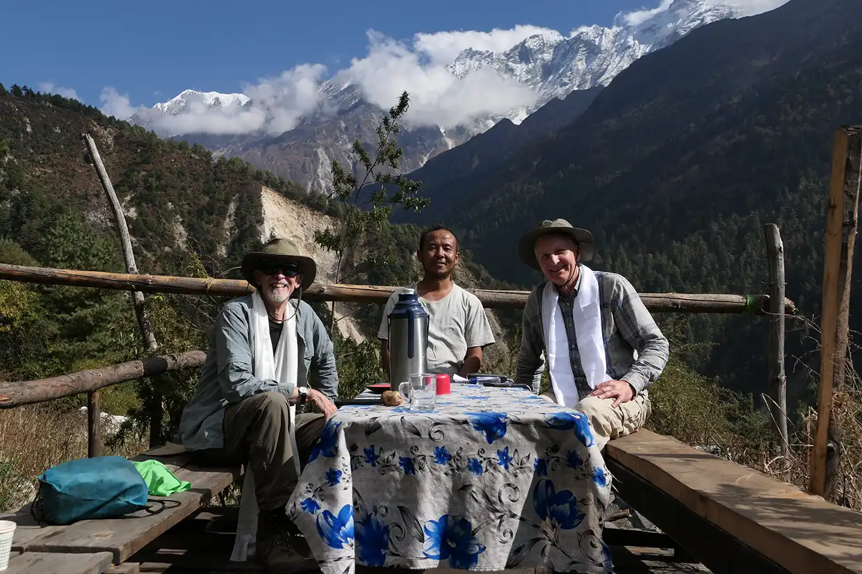 Two travelers and a local guide sit at an outdoor table at the teahouse in chhokamparo village.webp along the Manaslu Tsum Valley Trek.