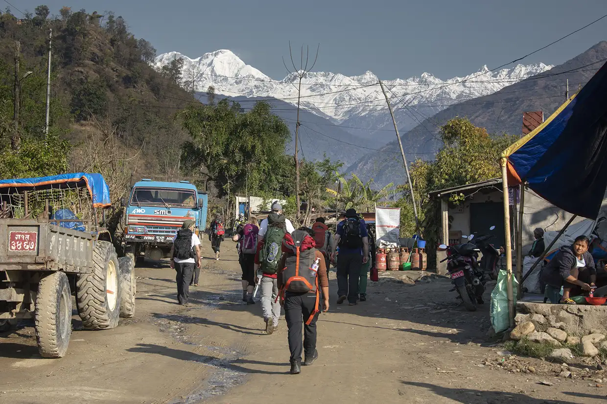 Trekkers with large backpacks walk down a dirt road in a mountain village, setting off on the scenic Nar Phu Valley Trek toward snow-capped peaks.