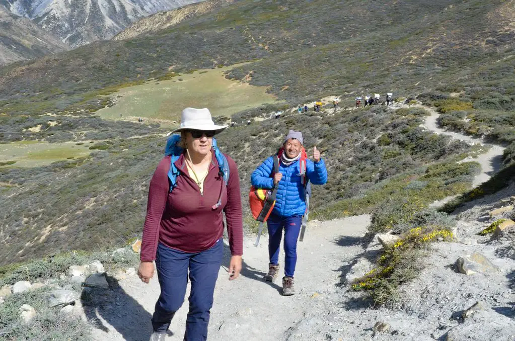 A female hiker and her local guide walk up a winding dirt path through rolling mountain hills during their Upper Dolpo Trek.
