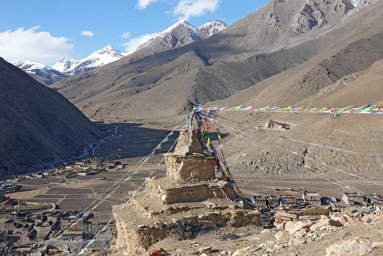 A stone chorten adorned with colorful prayer flags overlooks a sprawling mountain valley village and winding river during the iconic Upper Dolpo Trek.