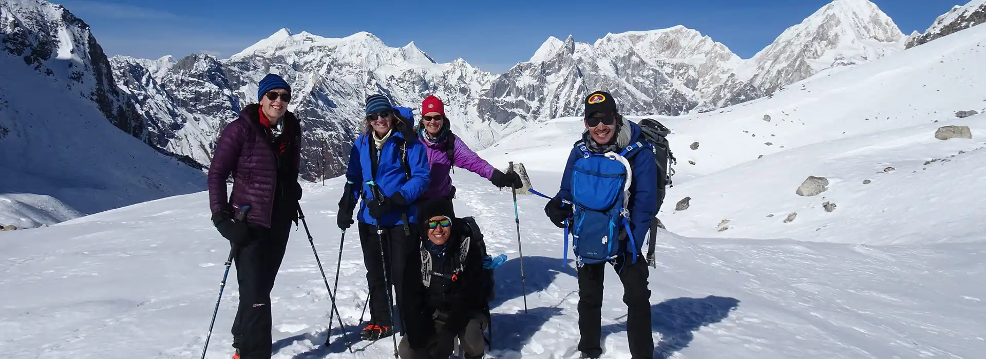 A cheerful group of five hikers poses together on a snowy high-altitude trail, surrounded by massive, jagged white peaks during their Nar Phu Valley Trek.