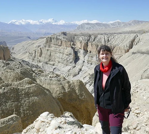 A female traveler smiles in front of a massive desert canyon and distant snow-covered Himalayan peaks during an Upper Mustang Jeep Tour.