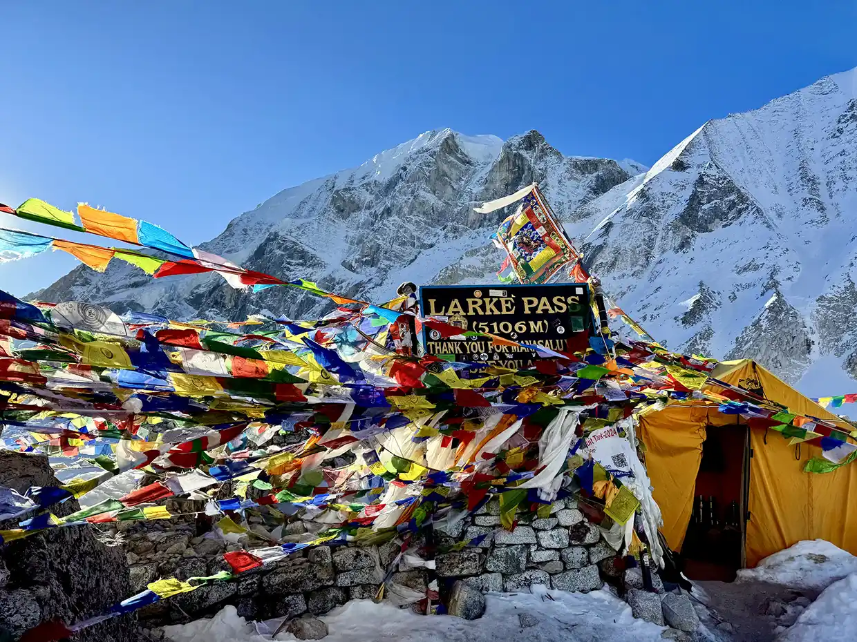 Colorful Buddhist prayer flags flutter around the Larke Pass signpost at 5,106 meters against a backdrop of snow-covered mountains on the Manaslu Tsum Valley Trek.