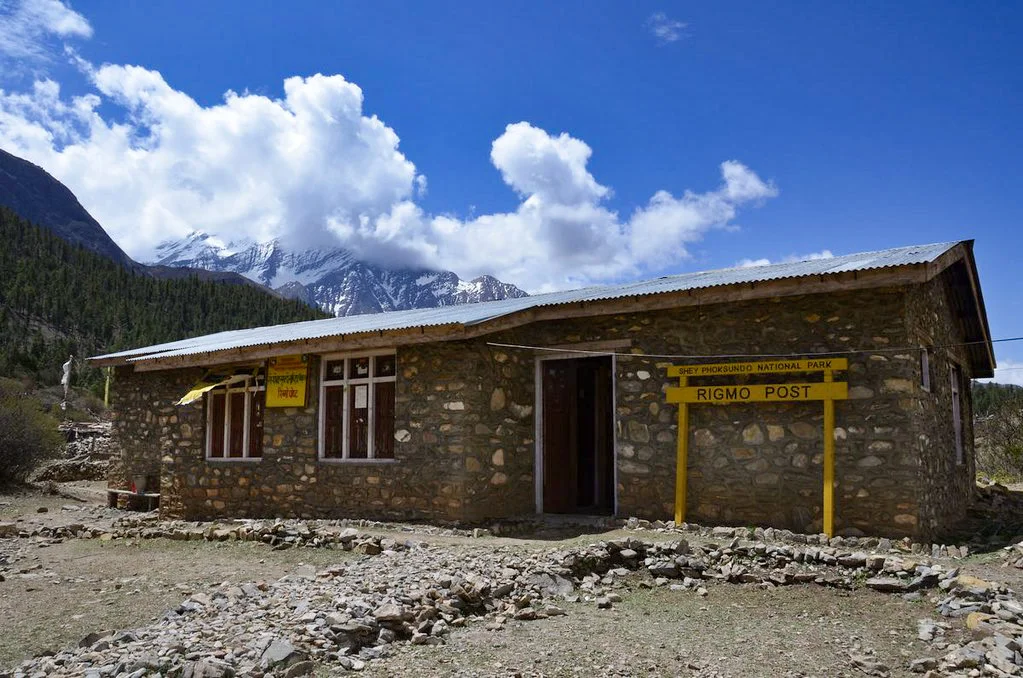 A stone building serves as the Rigmo Post checkpoint under a bright blue sky with snowy mountains in the background during an Upper Dolpo Trek.